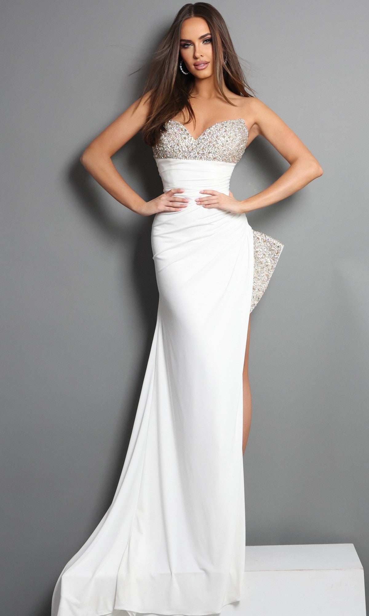 A woman poses against a gray wall wearing the Long Formal Dress: Jovani 49577 in Off White—a striking column gown featuring a strapless crepe silhouette, sequined bodice with side embellishment, thigh-high slit, and floor-length skirt. The crystal-embellished sweetheart neckline provides an extra touch of elegance to this formal look.